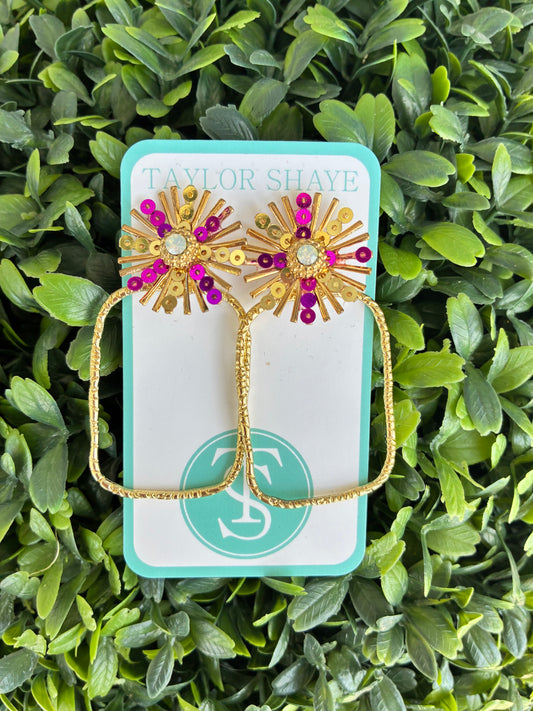 A pair of gold-toned starburst stud earrings with purple and gold rhinestone accents, displayed on a turquoise and white Taylor Shaye card against a background of green foliage.