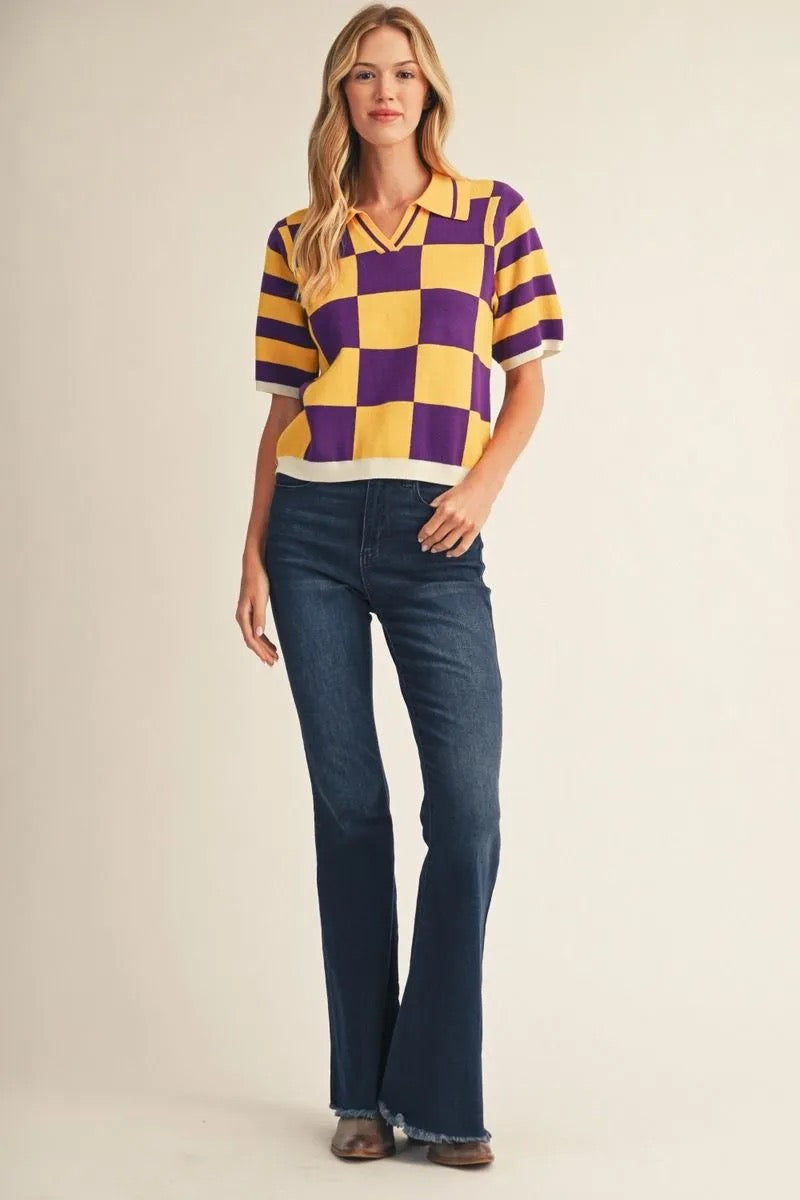 A woman modeling a short-sleeve knit polo shirt with a purple and yellow checkerboard pattern, paired with dark blue flared jeans and brown shoes, against a plain beige studio background.