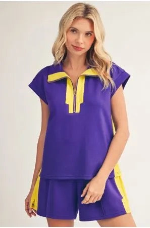 A woman modeling a purple short-sleeve top and matching shorts set with yellow contrast trim and side panels, photographed against a plain light background.