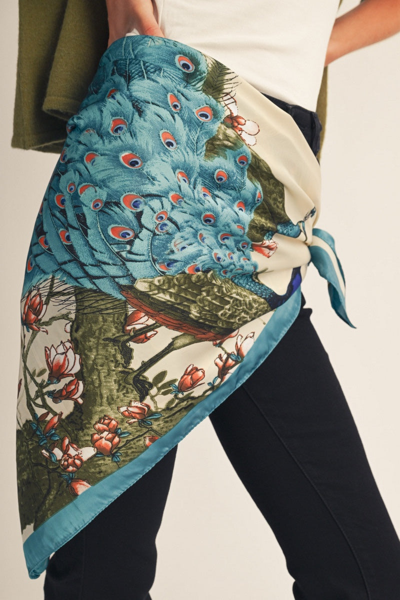 A person wearing a large square scarf tied around the waist, featuring a detailed print of blue peacock feathers and green foliage on a cream background.