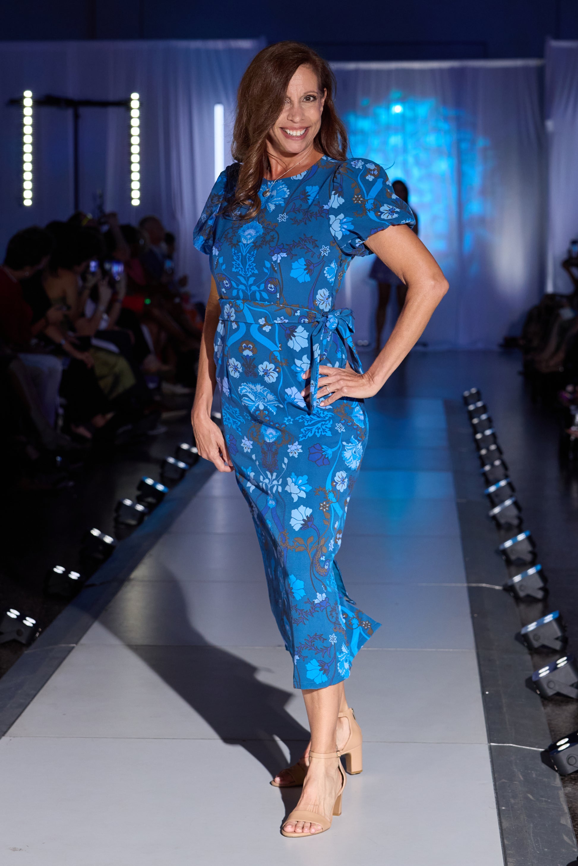 A woman models a blue floral midi dress with short puff sleeves and a tie waist, walking down a fashion runway with an audience in the background.
