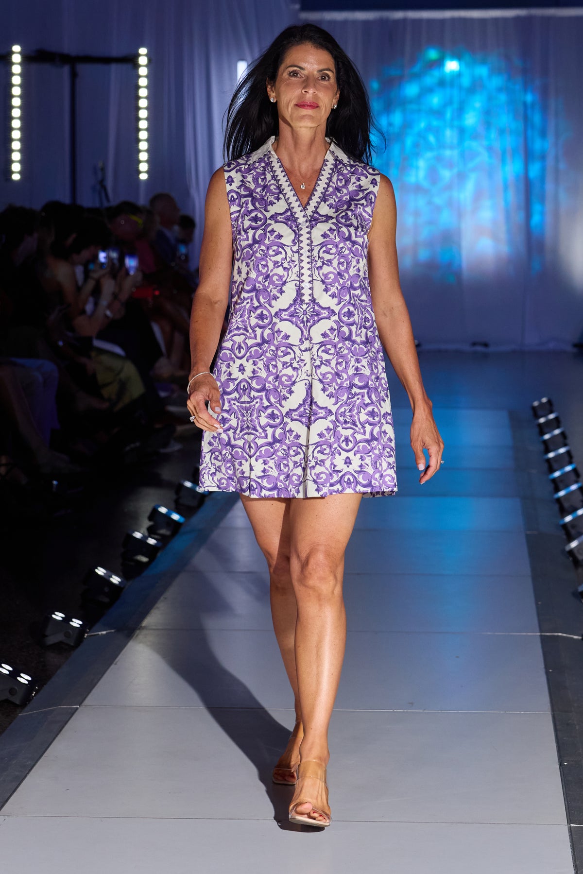 A woman walks down a fashion runway wearing a sleeveless purple and white patterned mini dress with a V-neckline 
