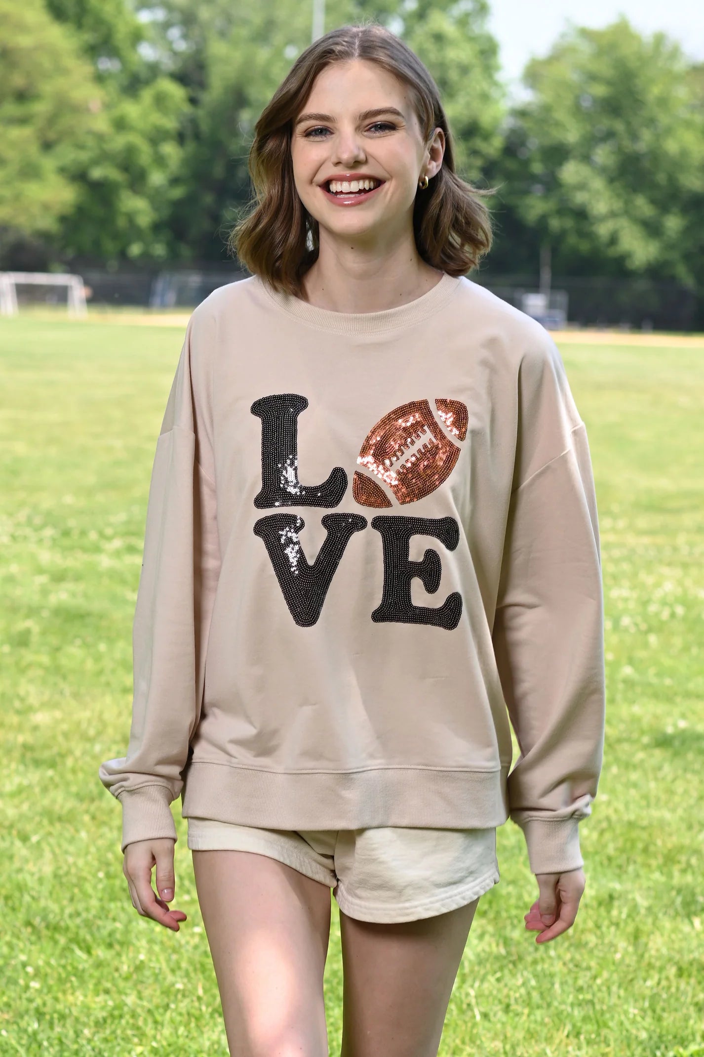 A smiling young woman standing outdoors on a grassy field wearing a beige crewneck sweatshirt with a 'LOVE' football sequin graphic 