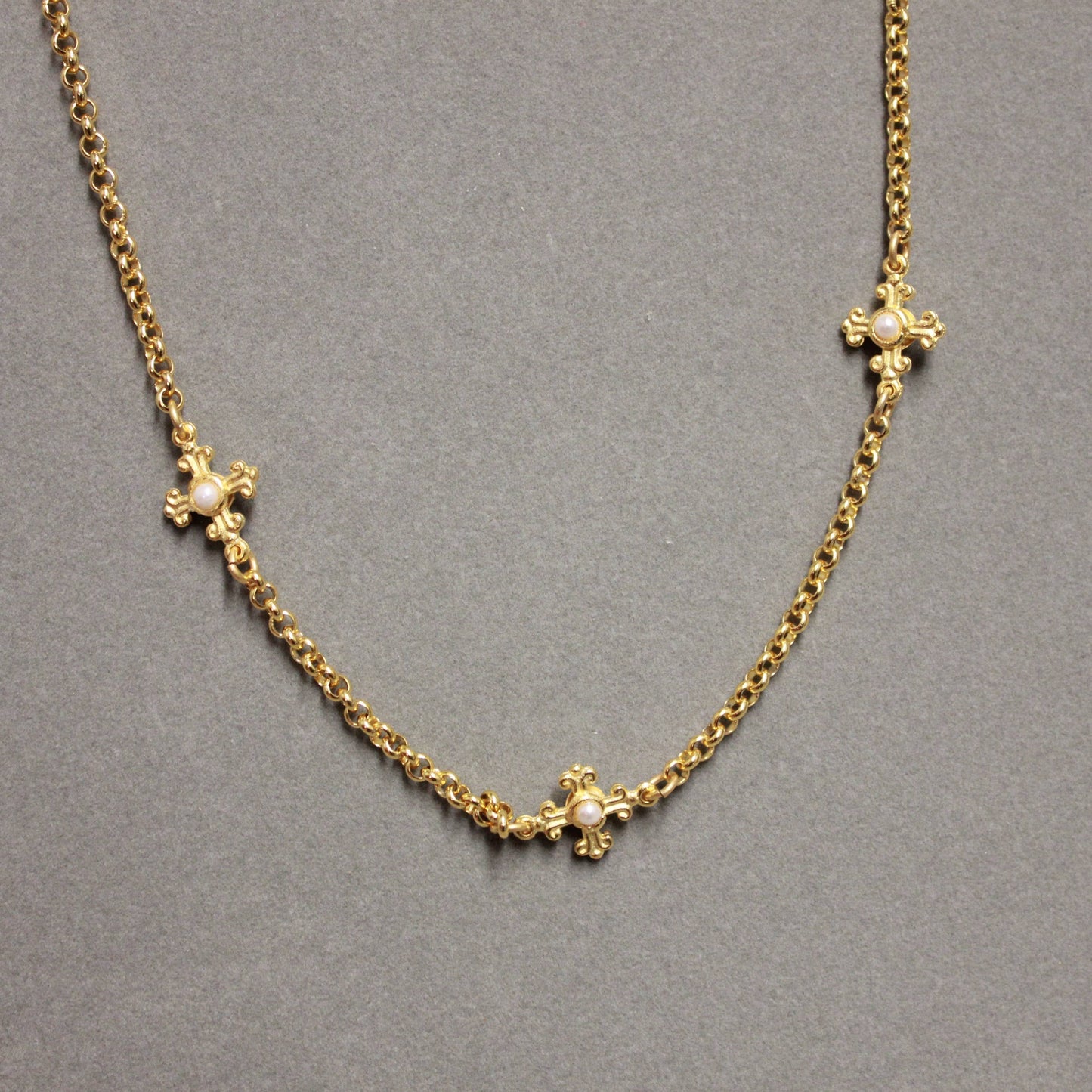A gold chain necklace with three ornate cross motifs, each adorned with a small clear stone, displayed on a neutral gray background.