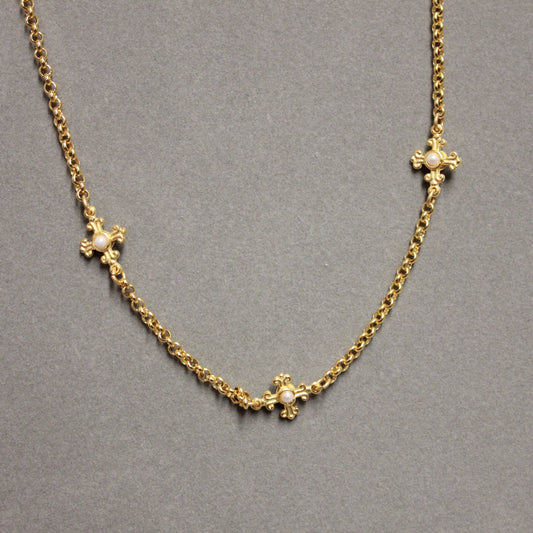 A gold chain necklace with three ornate cross motifs, each adorned with a small clear stone, displayed on a neutral gray background.