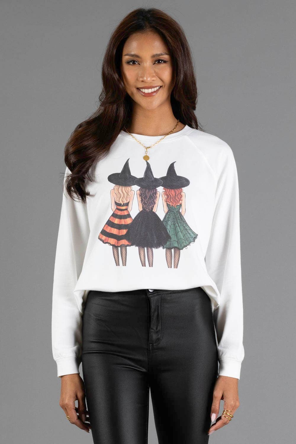 A woman modeling a white long-sleeve crewneck sweatshirt with a graphic of three illustrated witches wearing black hats and colorful dresses, paired with black pants, against a solid gray studio background.