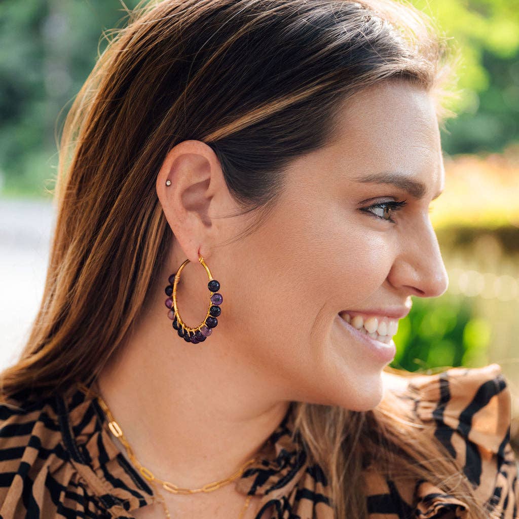 A close-up side profile of a smiling woman wearing a gold hoop earring adorned with dark purple and black beads, set against a soft, blurred outdoor background.