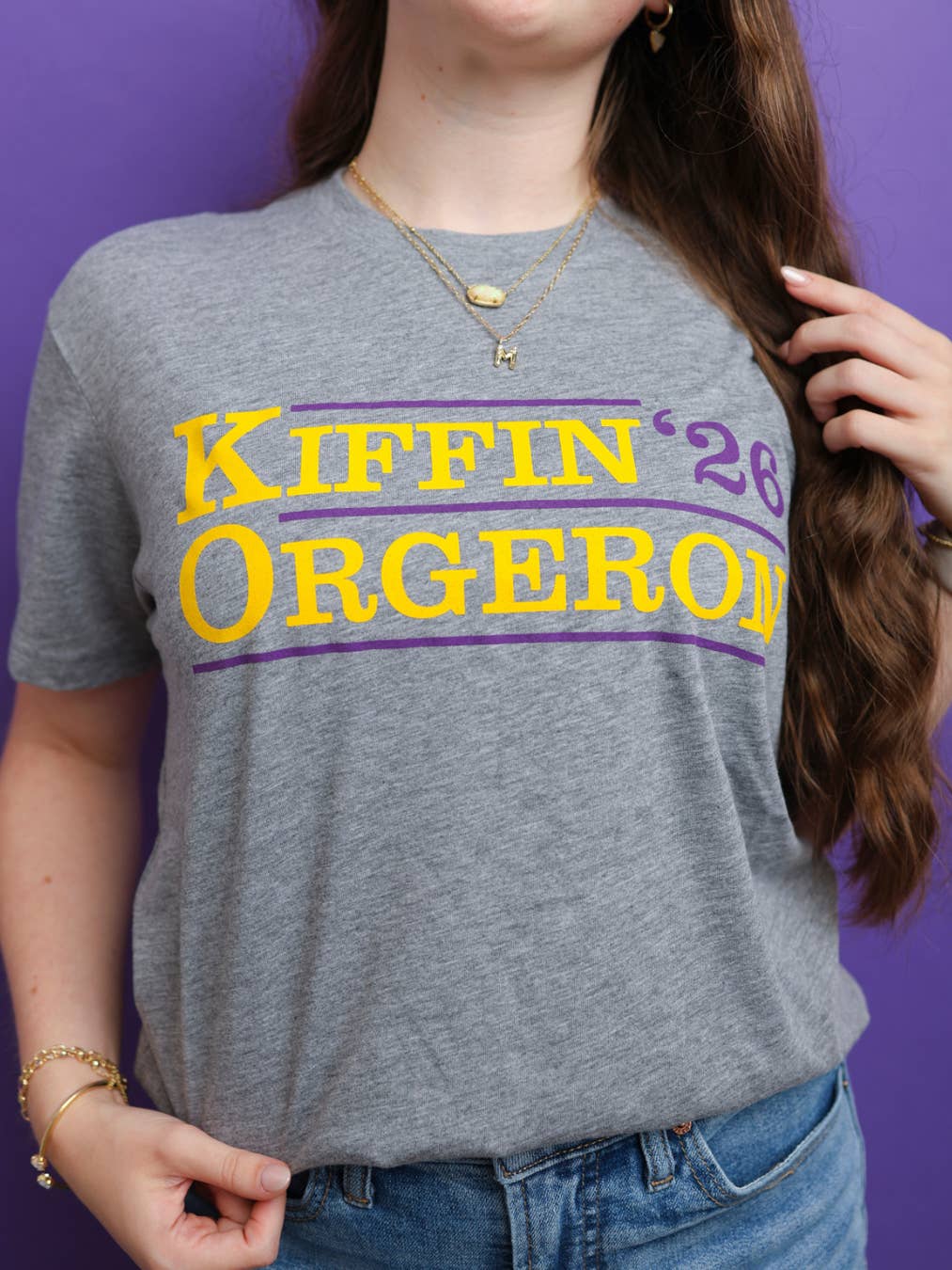 A woman wearing a heather gray graphic t-shirt with yellow and purple text reading 'Kiffin '26 ORGERON' against a solid purple background.