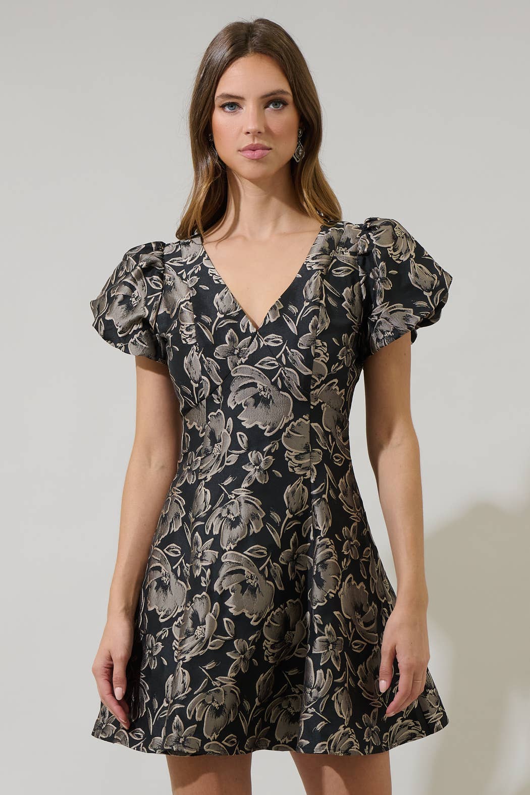 A woman modeling a short puff-sleeve V-neck dress with a black and beige floral pattern, set against a plain light gray studio background.