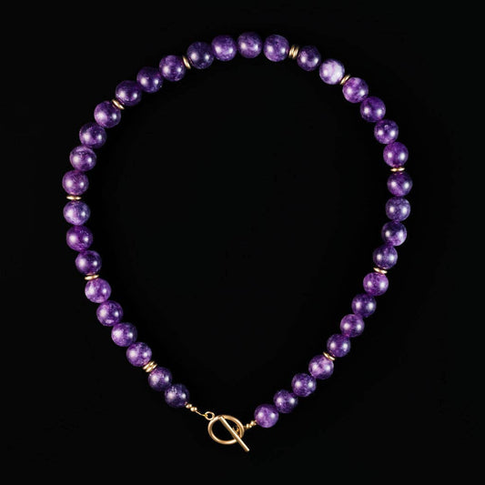 A beaded necklace made of round purple beads separated by small gold spacer beads, with a gold toggle clasp, displayed on a solid black background.