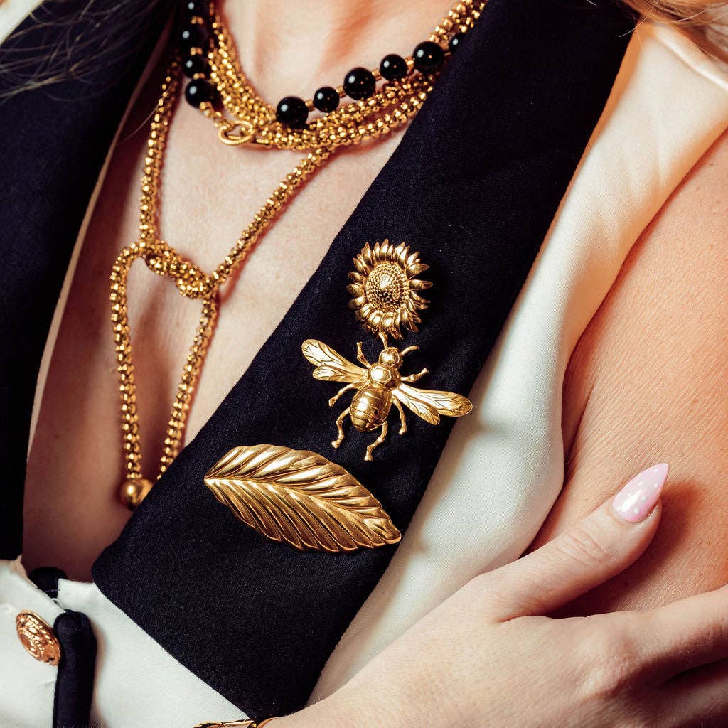 A close-up of a person wearing layered gold jewelry, including a prominent gold bee brooch with a sunflower detail, a gold leaf pin, and a beaded necklace over a black blazer and cream blouse.