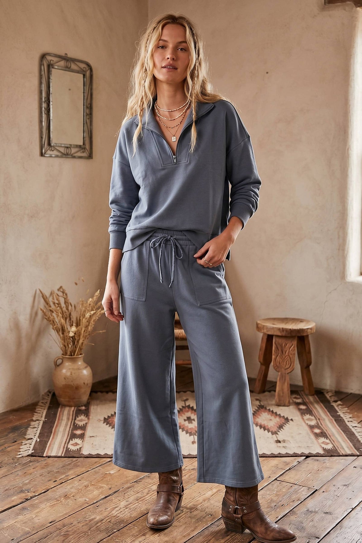 A woman stands in a rustic room wearing a matching dusty blue-gray lounge set consisting of a long-sleeve drawstring henley top and wide-leg drawstring pants, paired with brown leather boots.