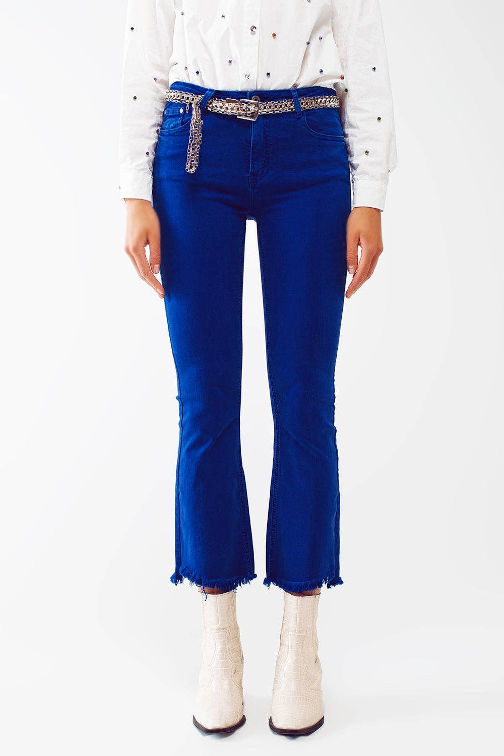 A model is shown from the waist down wearing bright royal blue cropped flare jeans with frayed hems, styled with a silver studded belt, a white embroidered long-sleeve top, and white ankle boots.