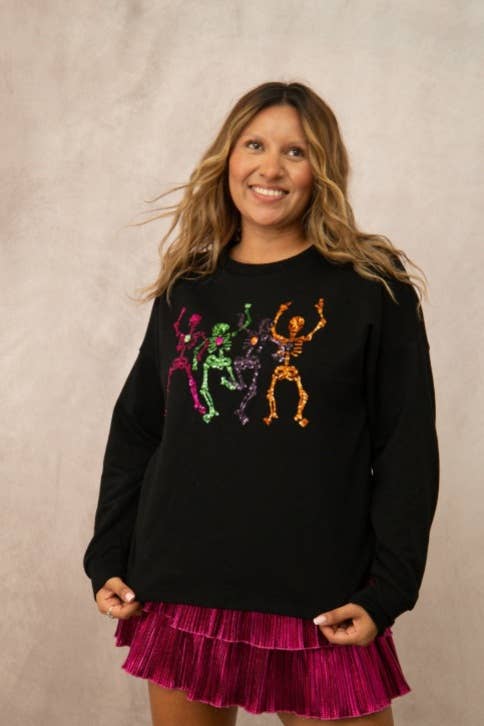 A woman modeling a black long-sleeve crewneck sweatshirt featuring a colorful graphic of three dancing skeletons in pink, green, and orange, paired with a magenta pleated skirt, against a neutral textured beige backdrop.