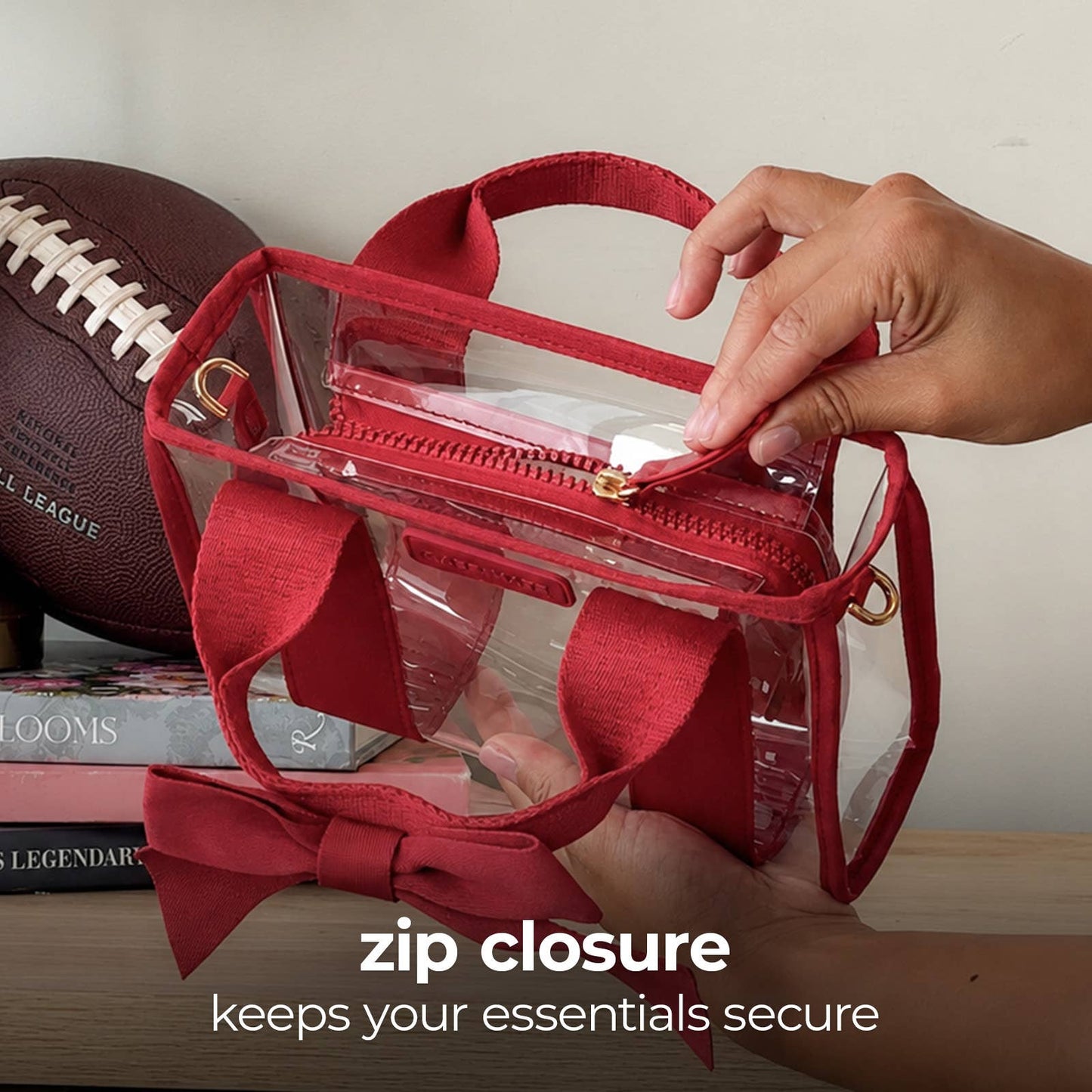 A clear plastic zippered pouch bag with red fabric handles, trim, and bow details, shown with a hand opening the top zipper. A football and books sit in the background, and text at the bottom reads 'zip closure keeps your essentials secure'.