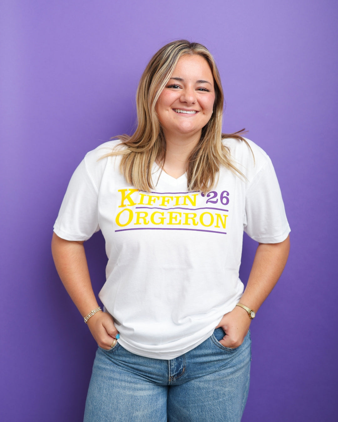 A smiling woman with blonde hair stands against a solid purple background wearing a white V-neck t-shirt with 'Kiffin '26 Orgeron' printed in yellow and purple across the chest, paired with blue jeans.