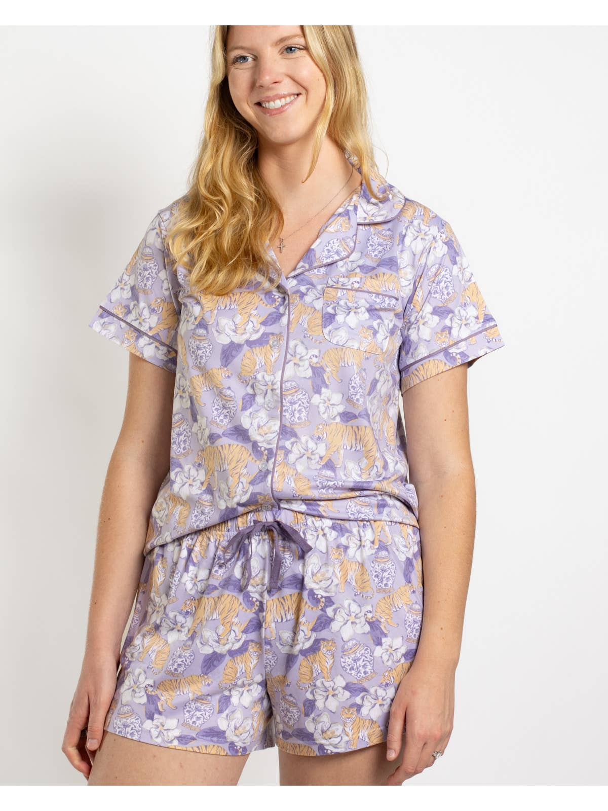A woman wearing a short-sleeve pajama set in lavender lavender with tigers, magnolias, and vests  featuring a button-up top and matching drawstring shorts, against a plain white background.