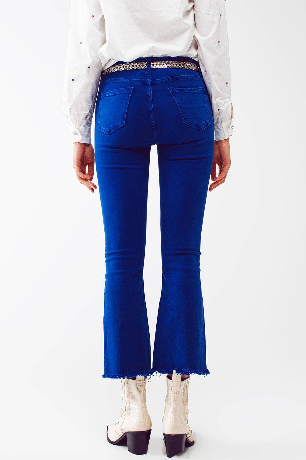 Rear view of a person wearing bright royal blue flared jeans with a frayed hem, paired with a studded belt, white blouse, and white heeled boots against a plain white studio background.