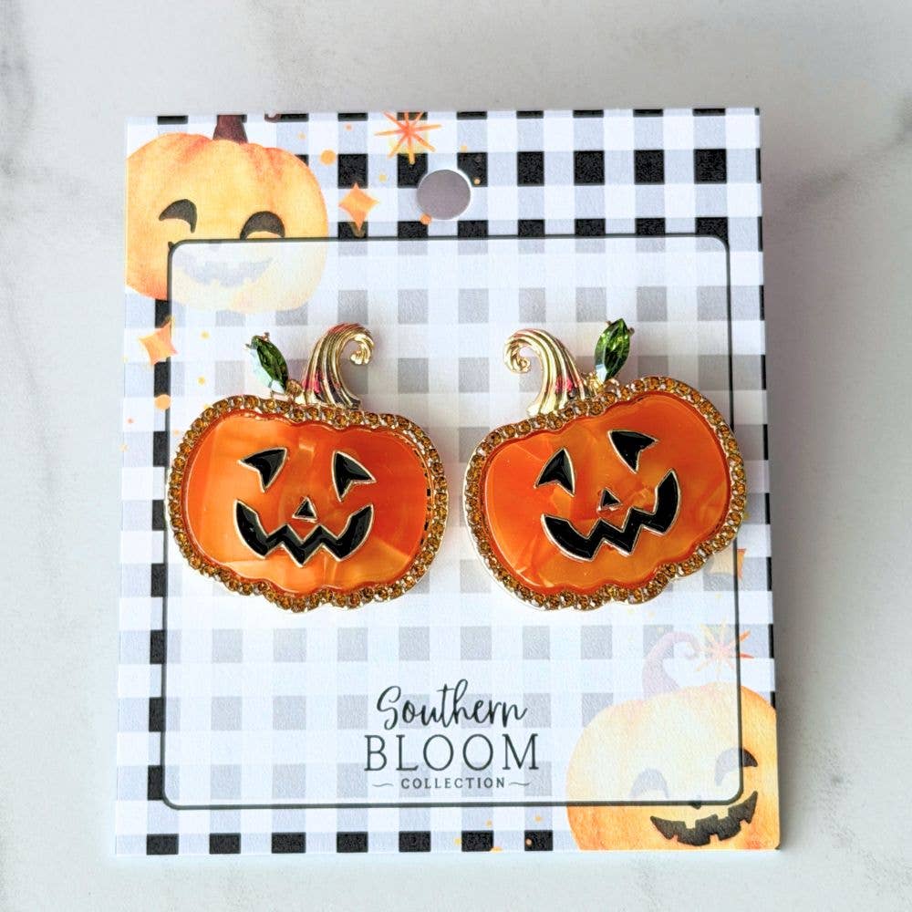 A pair of pumpkin-shaped earrings displayed on a branded card. The earrings are orange with black jack-o'-lantern faces and gold glittery scalloped edges, with small green stems.