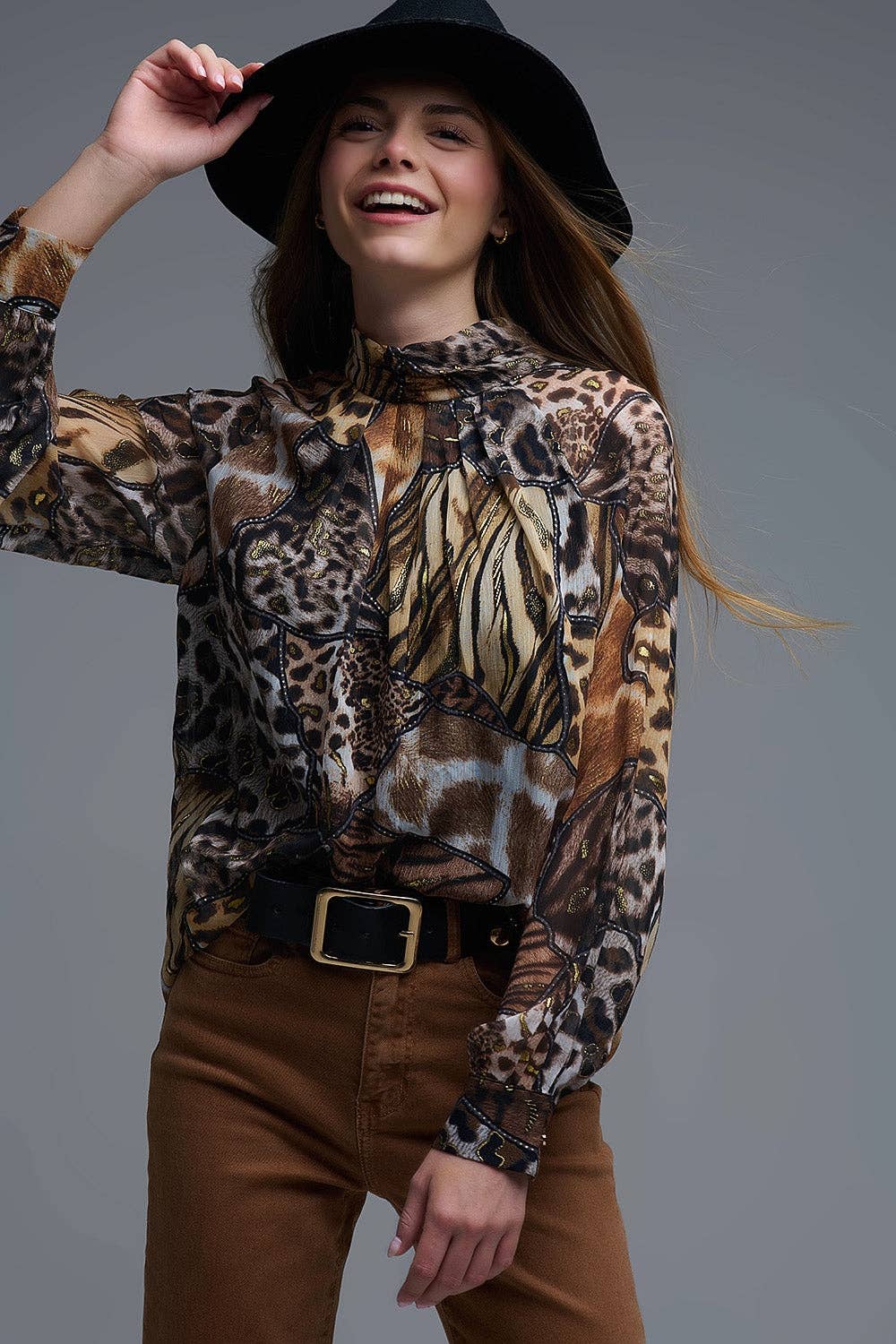 A woman wearing a long-sleeve high-neck blouse with an abstract animal print pattern in brown, beige, black, and white, styled with brown pants, a black belt, and a black wide-brim hat against a solid gray studio background.