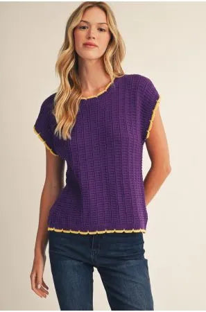 A woman modeling a purple short-sleeve knit top with yellow trim, paired with dark blue jeans, against a plain beige background.