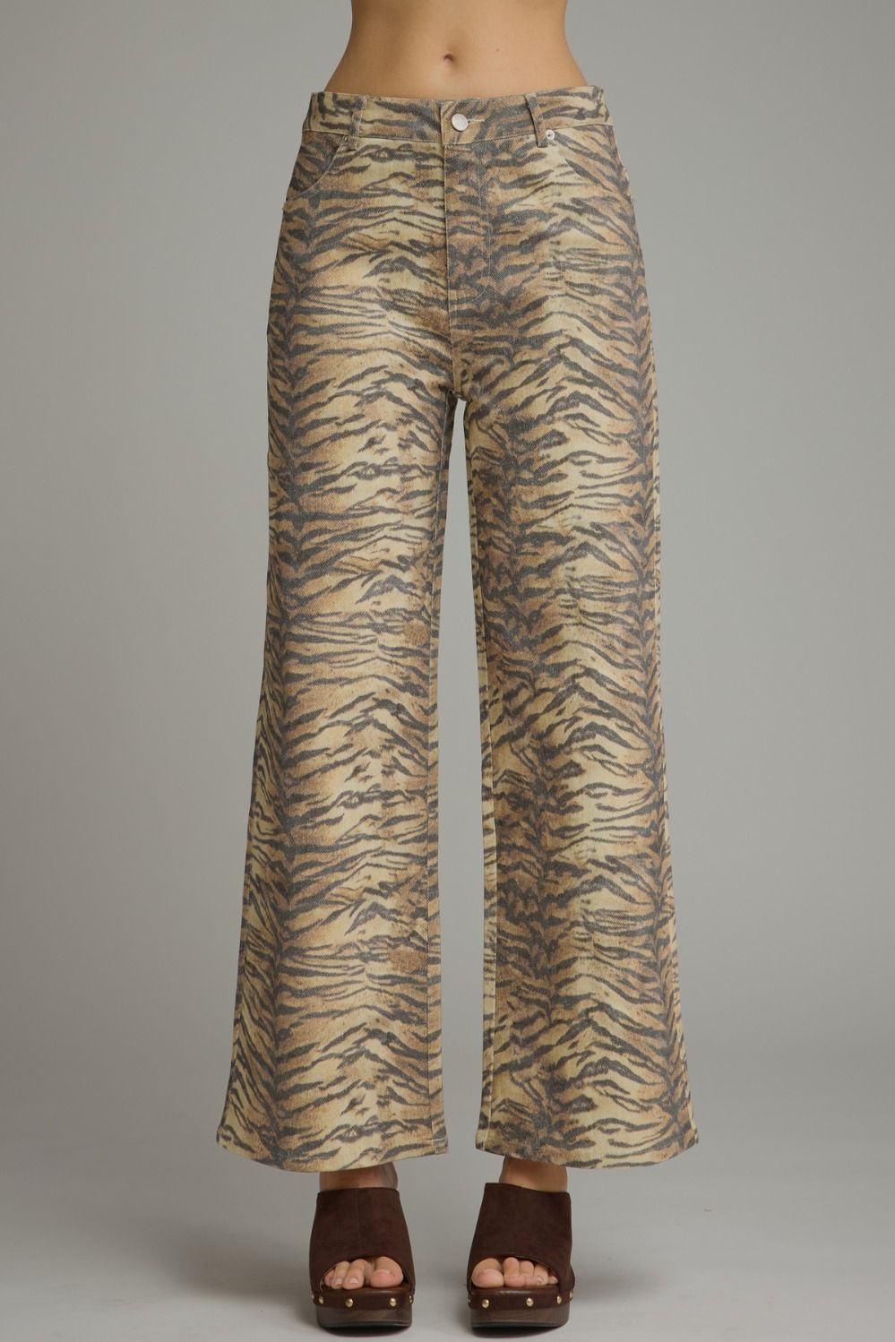 A model is shown from the waist down wearing high-waisted, wide-leg trousers with a beige and brown tiger-stripe pattern, paired with dark brown platform sandals, against a solid gray studio background.