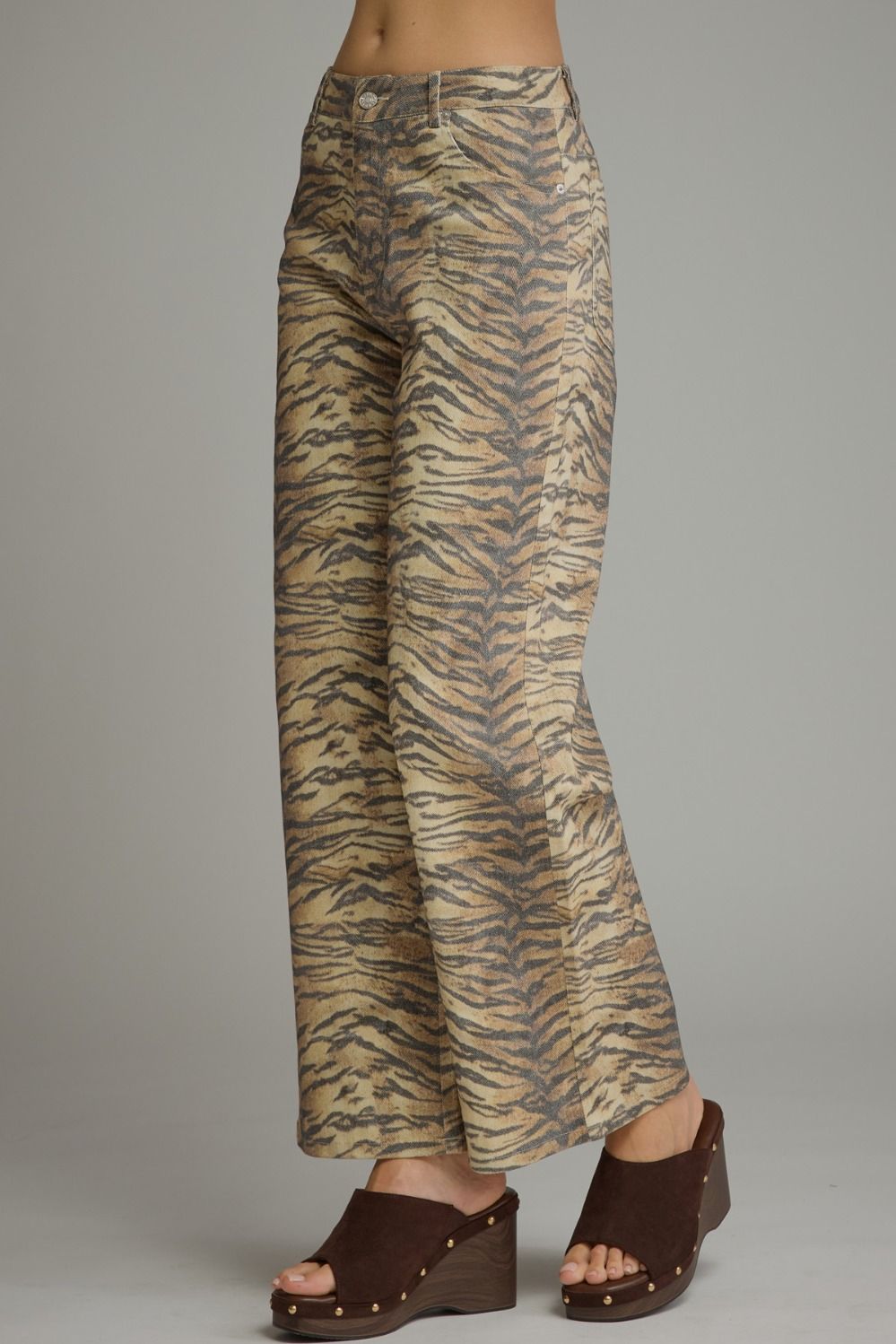 A person wearing wide-leg cropped pants with a beige and brown tiger stripe pattern, paired with dark brown wedge sandals, shown against a plain gray studio background.