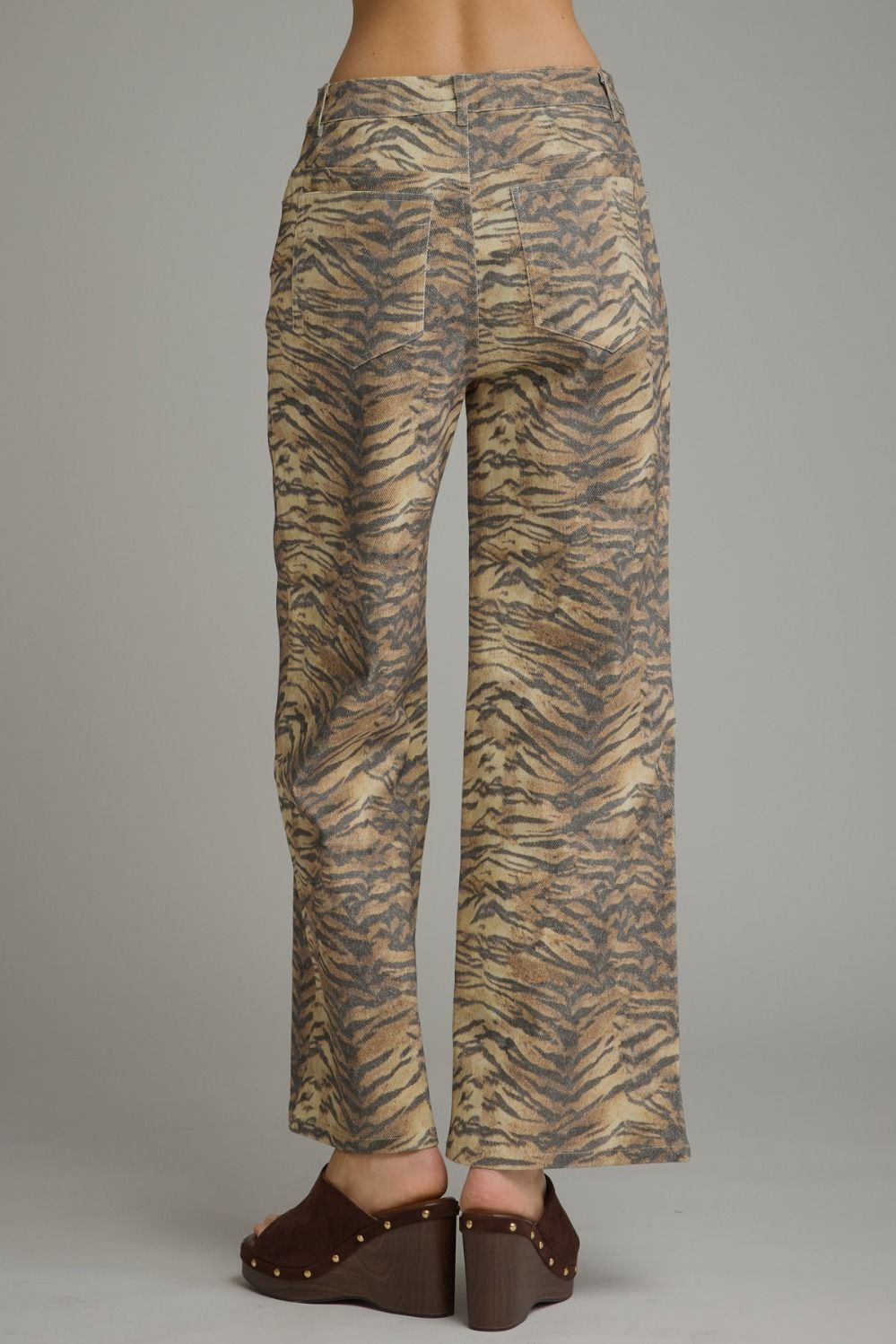 Wide-leg trousers with a beige and brown tiger stripe pattern, shown from the back on a model wearing brown wedge sandals against a neutral gray background.