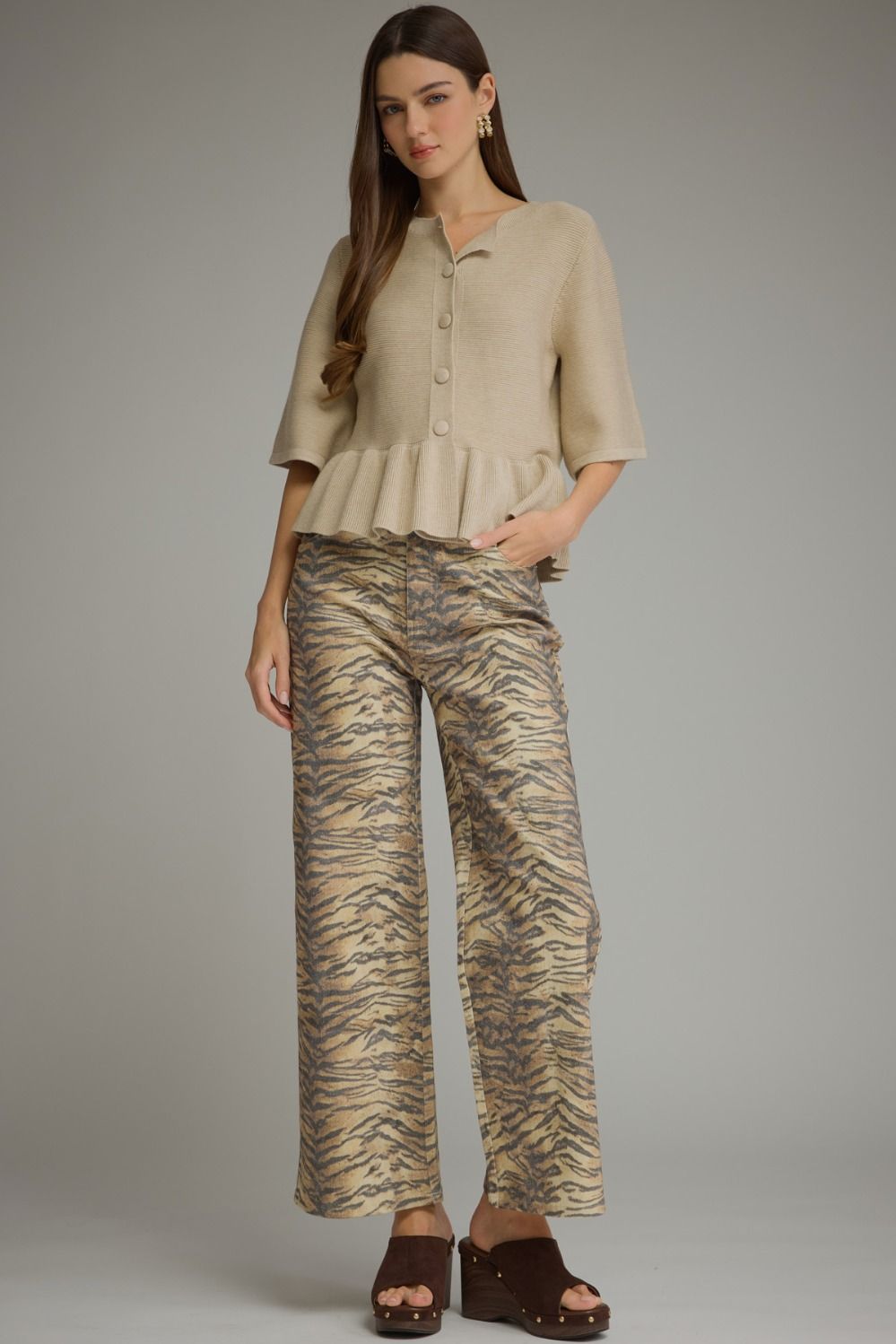 A woman modeling a beige short-sleeve peplum-style blouse and wide-leg trousers with a tan and brown tiger-stripe pattern, paired with dark brown slide sandals, against a solid gray studio background.