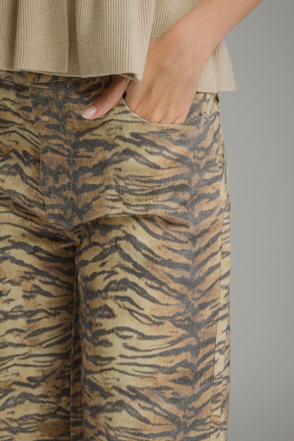 Close-up of a person wearing wide-leg trousers with a beige and brown tiger stripe pattern, paired with a ribbed beige top, against a solid gray background.