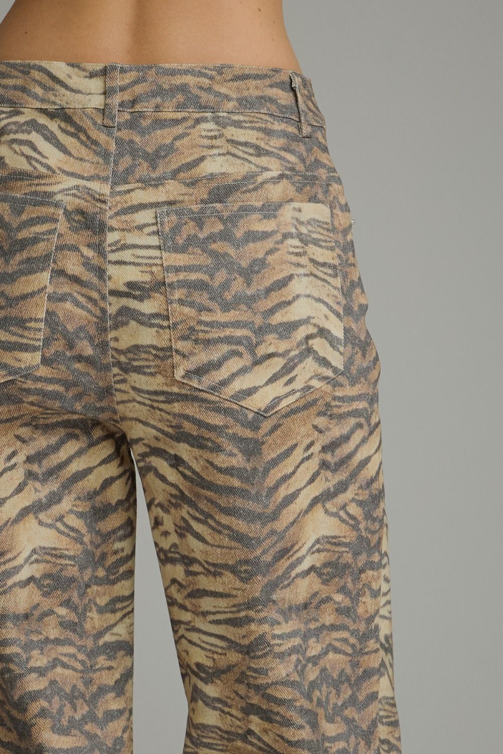 Close-up back view of a person wearing tiger-print pants with a beige, brown, and gray animal pattern, showing the back pockets and waistband against a plain gray background.