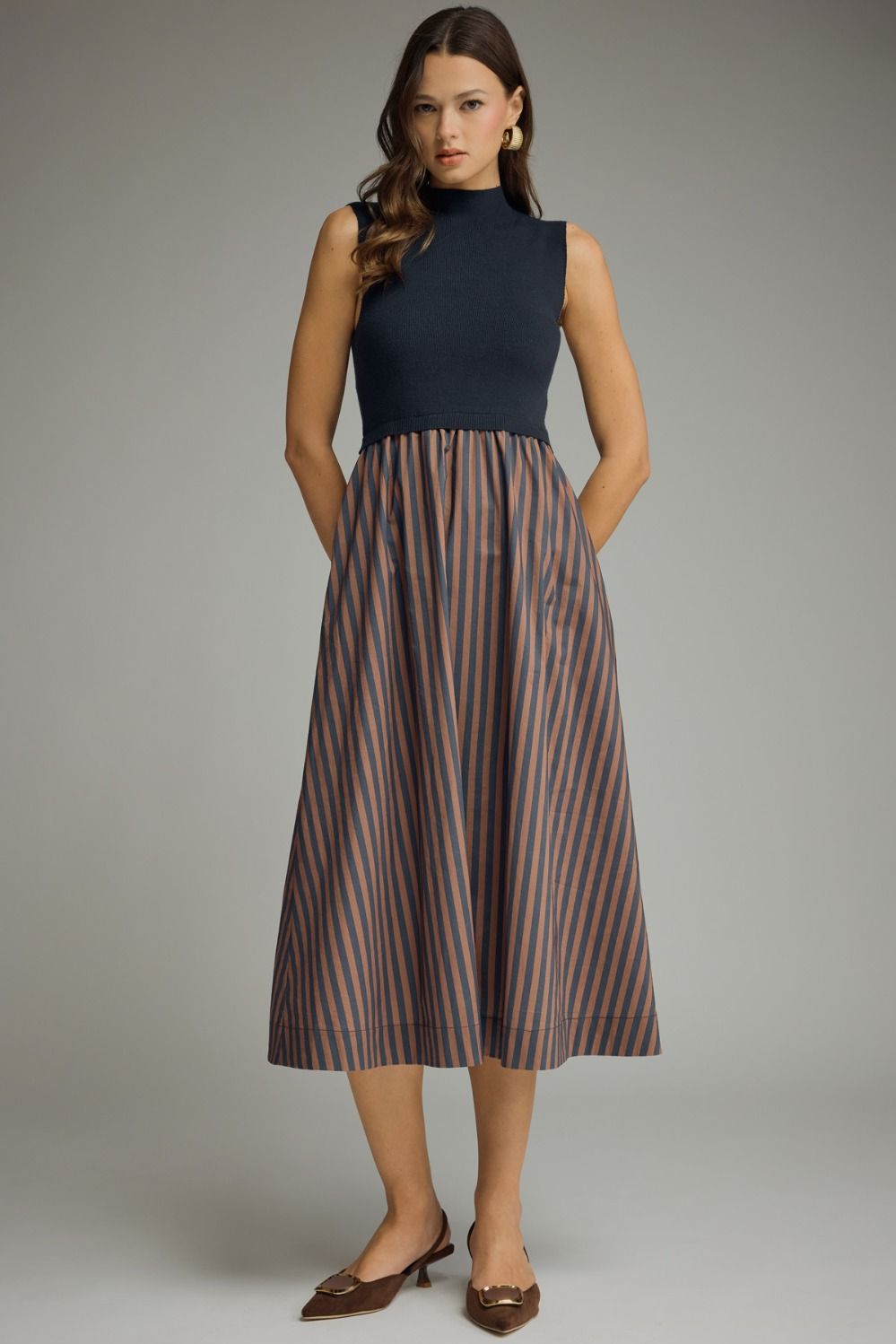 Woman wearing a sleeveless dress with a Navy top and a Brown and Navy striped skirt on a gray background