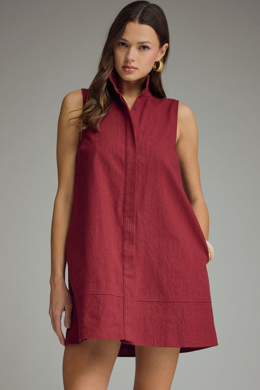 A woman modeling a sleeveless, high-neck, button-front linen dress in a deep burgundy red, set against a solid gray studio background.