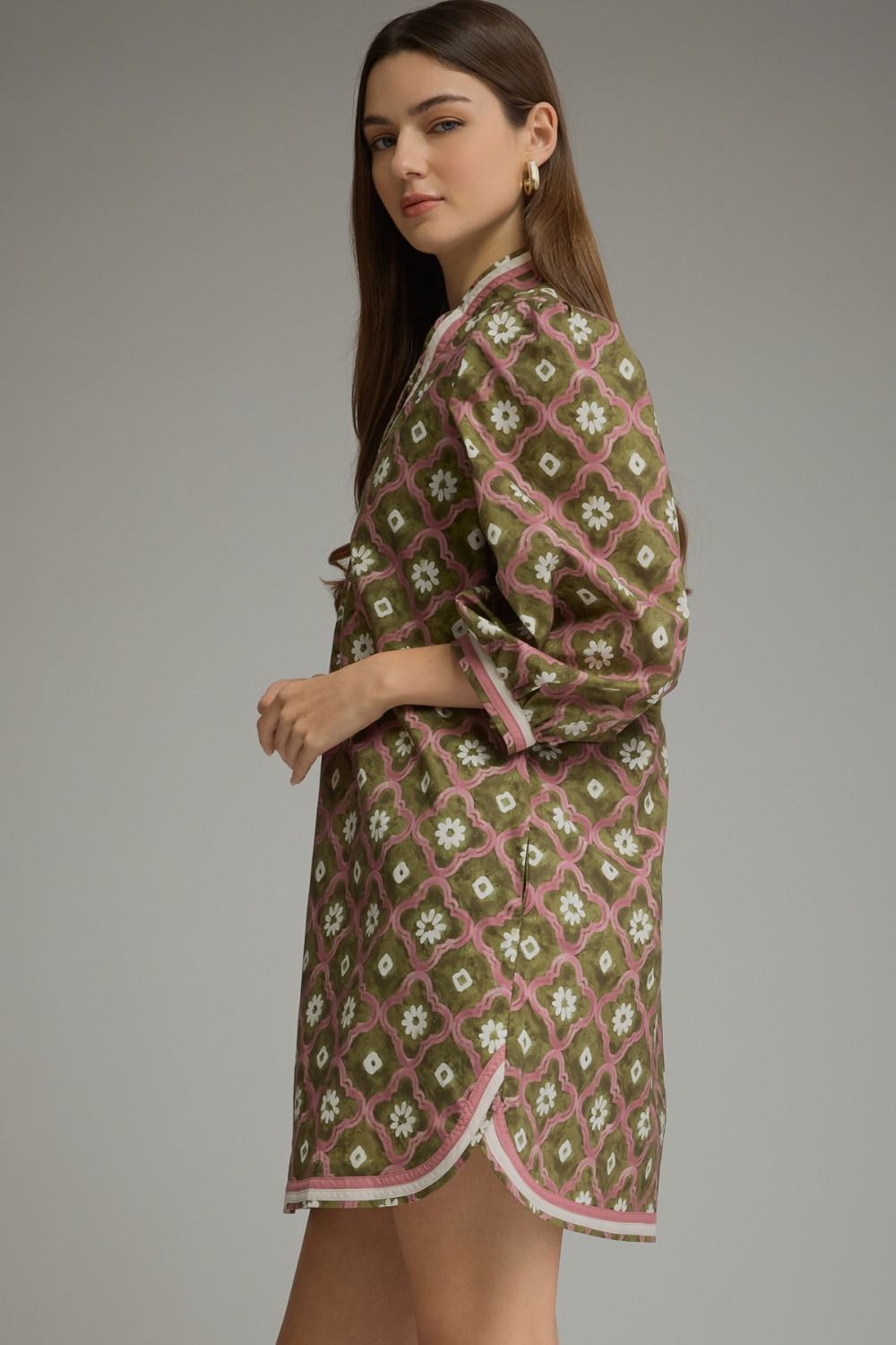 A woman modeling a short printed dress in olive green, pink, and white, shown from a side-back angle against a neutral gray studio background.