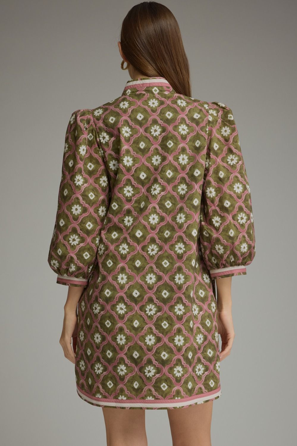 A woman shown from the back wearing a short puff-sleeve dress with an olive green and dusty pink geometric lattice pattern and white floral details, set against a solid gray studio background.