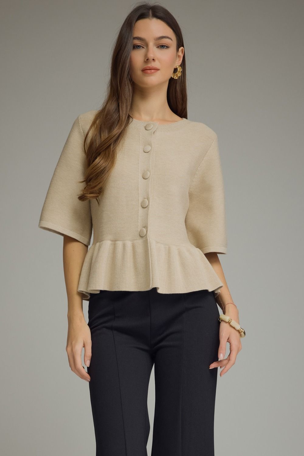 A woman modeling a beige short-sleeve cropped cardigan  with a buttoned front and peplum hem, paired with dark trousers, against a plain gray studio background.