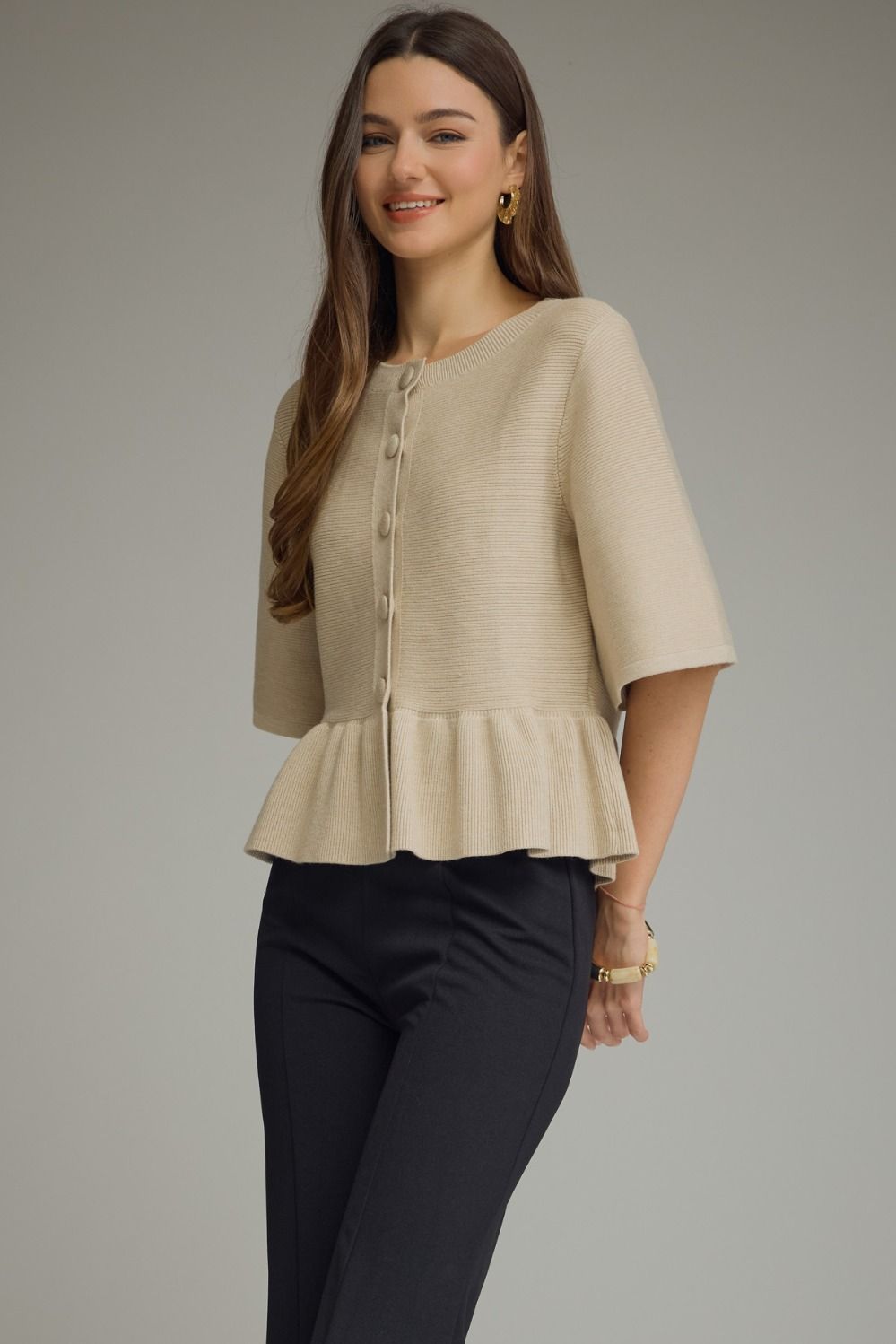 A woman modeling a beige short-sleeve knit peplum top with a button-front detail, paired with dark pants, against a neutral gray studio background.