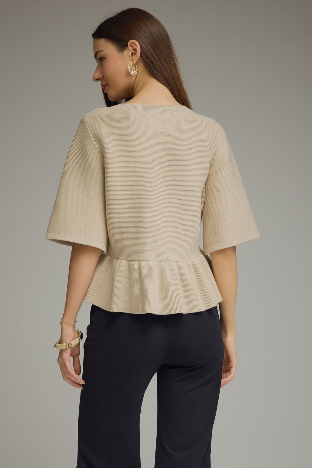 A woman shown from the back wearing a light beige short-sleeve knit top with a peplum hem and dark navy pants, against a plain gray studio background.