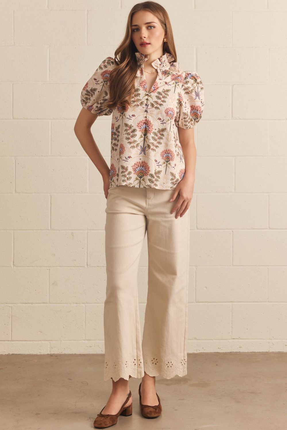 A woman modeling a cream puff-sleeve blouse with a pink and green floral print, paired with cream cropped pants and brown shoes, standing against a light beige brick wall.