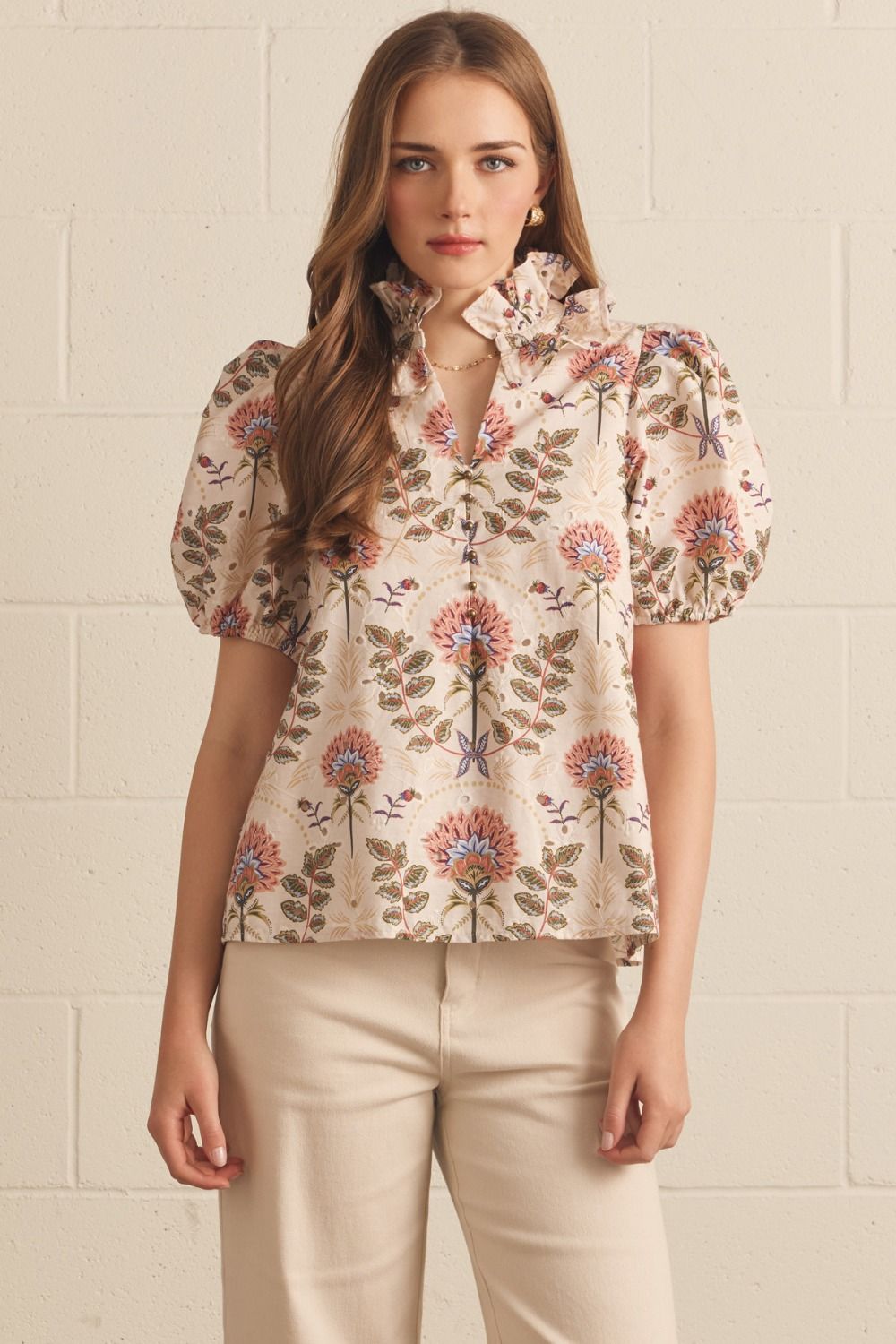 A woman wearing a beige puff-sleeve blouse with a colorful floral print, paired with beige pants, standing against a light beige brick wall.