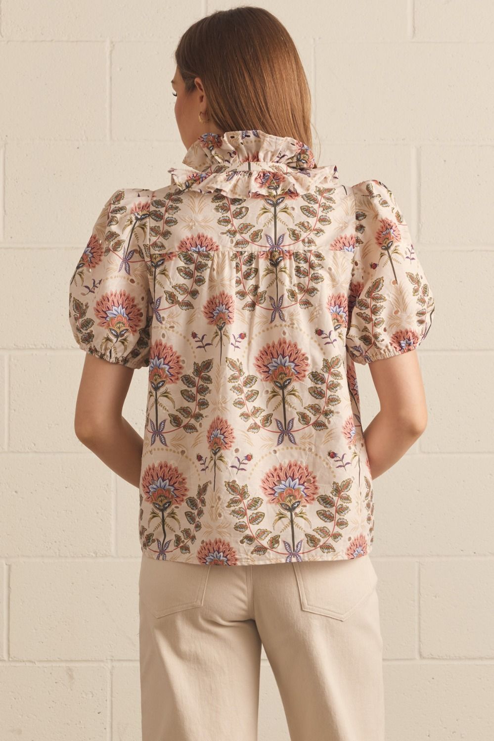 A woman viewed from behind wearing a cream-colored blouse with a colorful floral pattern, paired with beige trousers, standing against a light brick wall.
