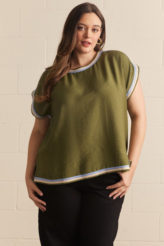 A woman modeling a short-sleeve olive green top with white and gray striped trim on the collar, sleeve cuffs, and hem, paired with black pants against a light beige brick wall.