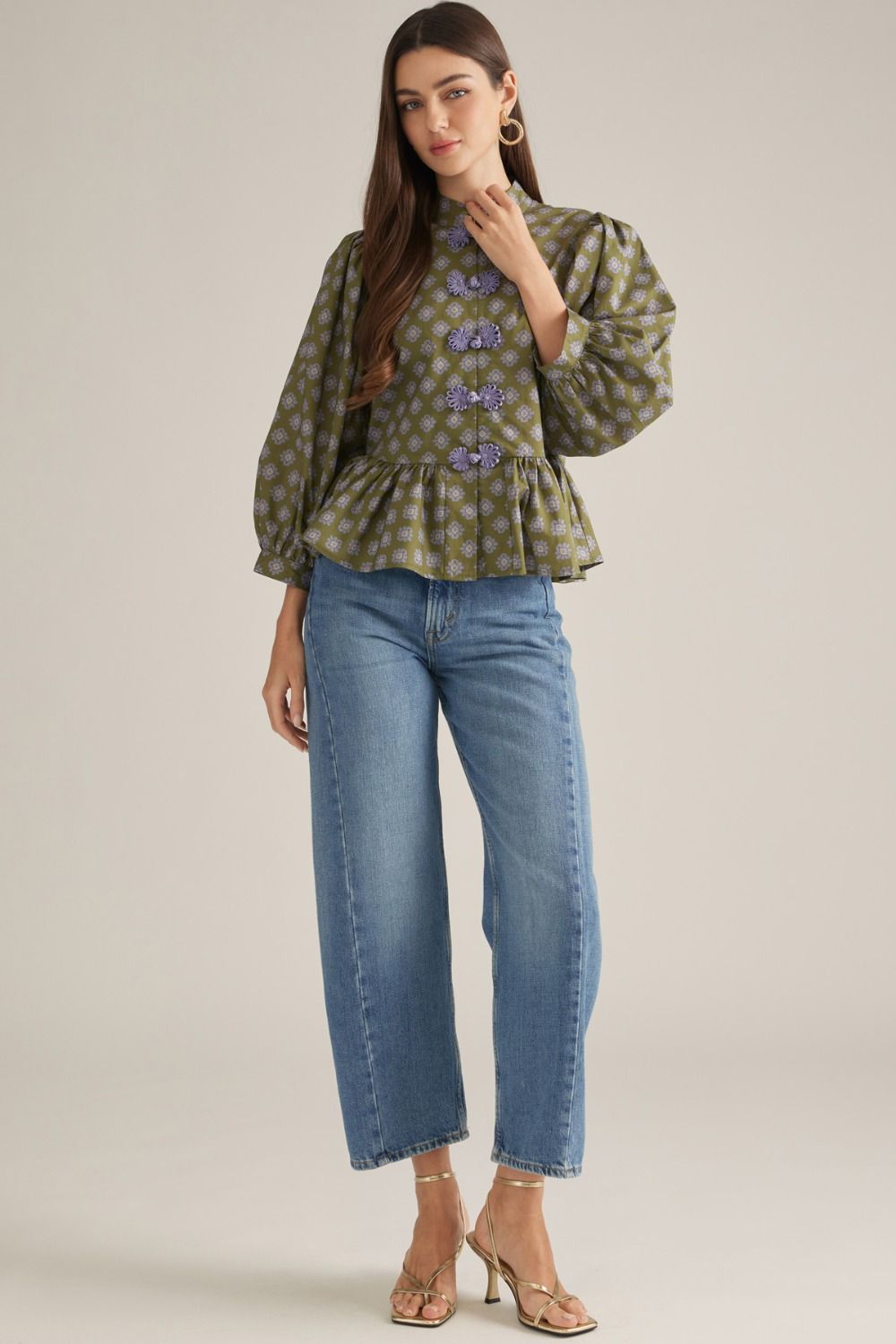 A woman models a green and gold patterned blouse with lavender decorative buttons, paired with blue jeans and gold heeled sandals, against a neutral studio background.