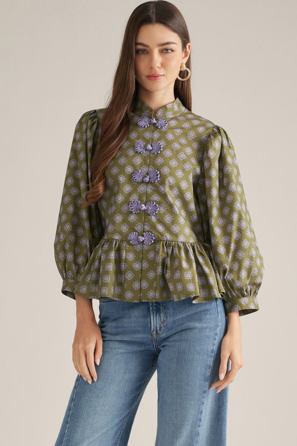 A woman modeling a green patterned blouse with lavender floral button details and blue jeans, standing against a plain beige studio background.