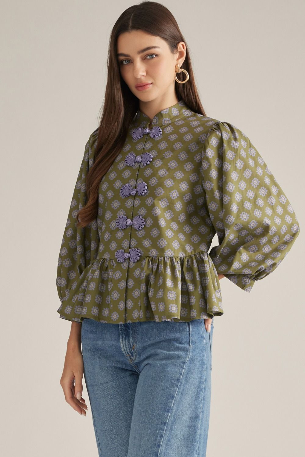 A woman modeling a green patterned blouse with lavender floral embroidery and decorative buttons, paired with blue jeans, against a plain beige studio background.