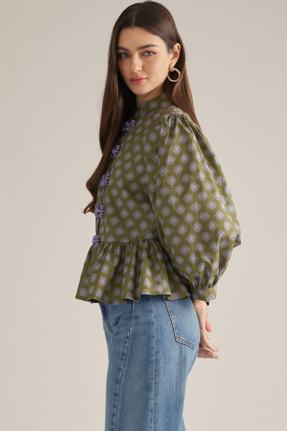 A woman modeling a green patterned blouse with lavender floral accents and blue jeans against a plain beige studio background.