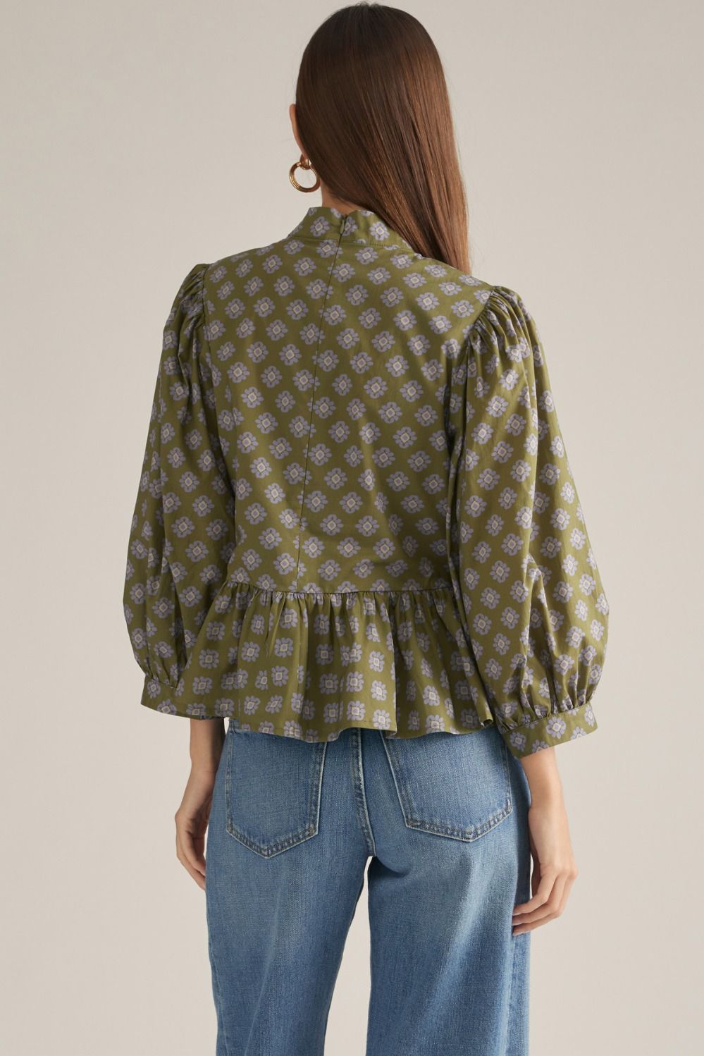 A woman shown from the back wearing a sage green long-sleeve blouse with a small white geometric pattern, puffed shoulders, and a ruffled hem, paired with blue jeans against a light beige background.