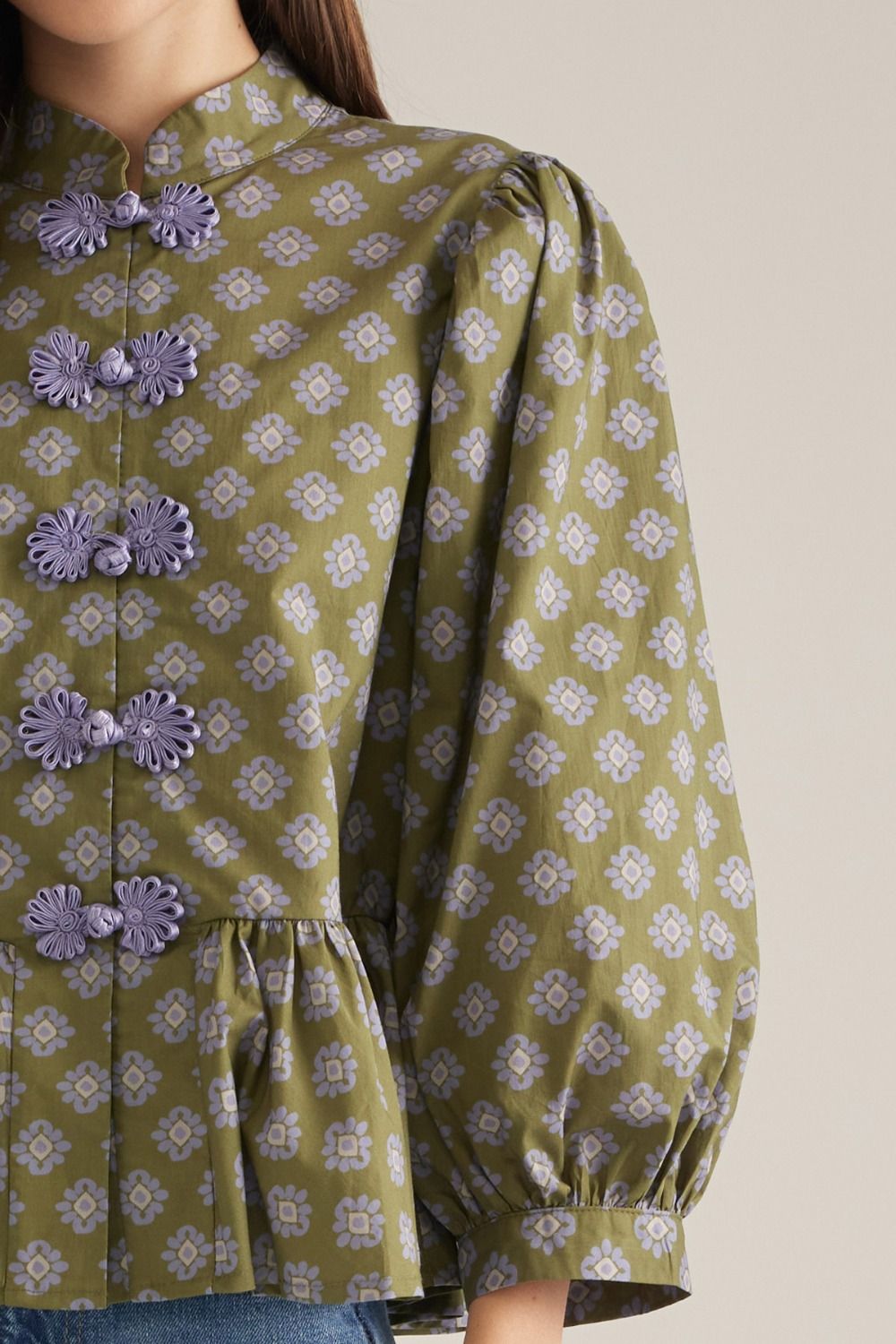 Close-up of a woman wearing a green long-sleeve blouse with a repeating floral pattern, lavender decorative frog closures, and a gathered peplum hem, paired with blue jeans against a plain beige background.