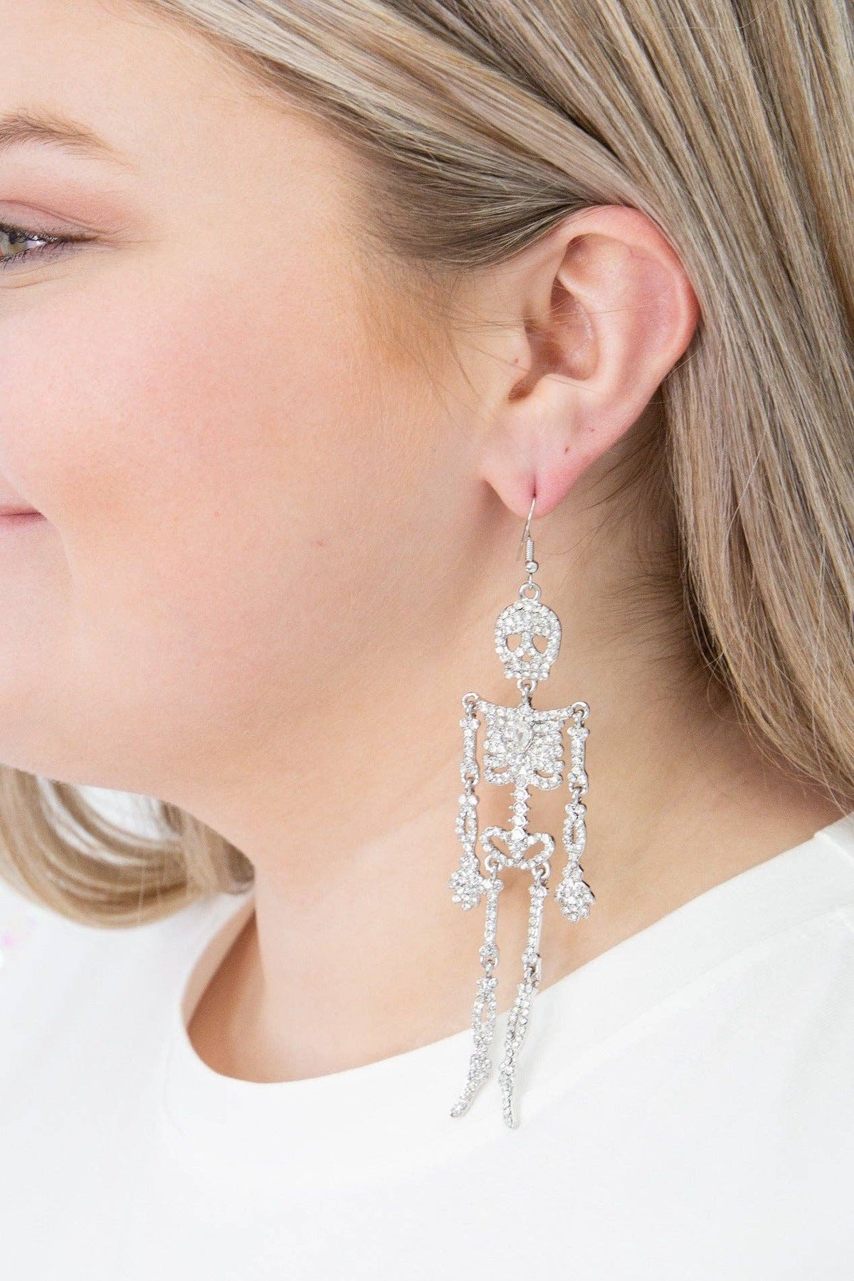 A close-up of a woman wearing a long, dangling skeleton-shaped earring made of sparkling clear stones on a silver-tone base.