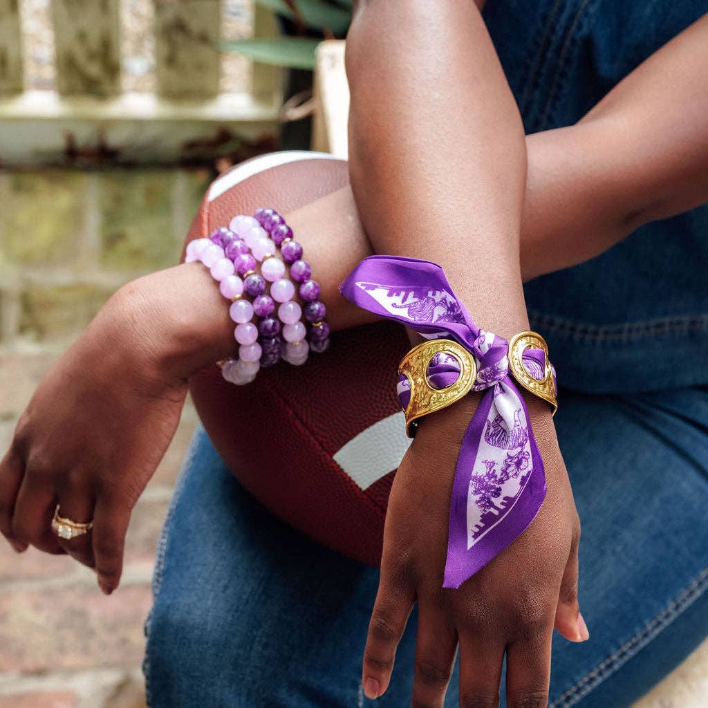 A close-up lifestyle shot of a person's arms and hands, showcasing a purple and white patterned fabric wrist scarf tied in a bow with gold-toned hardware, alongside purple and white beaded bracelets.
